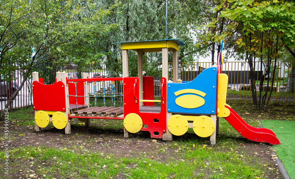 A wooden train of bright red, blue and yellow colors against a background of green trees and residential buildings on a clear sunny day. Playgrounds, sports, health entertainment.