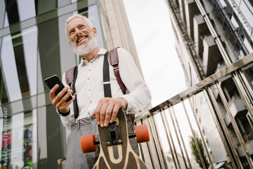 Grey mature man using cellphone while standing with skateboard
