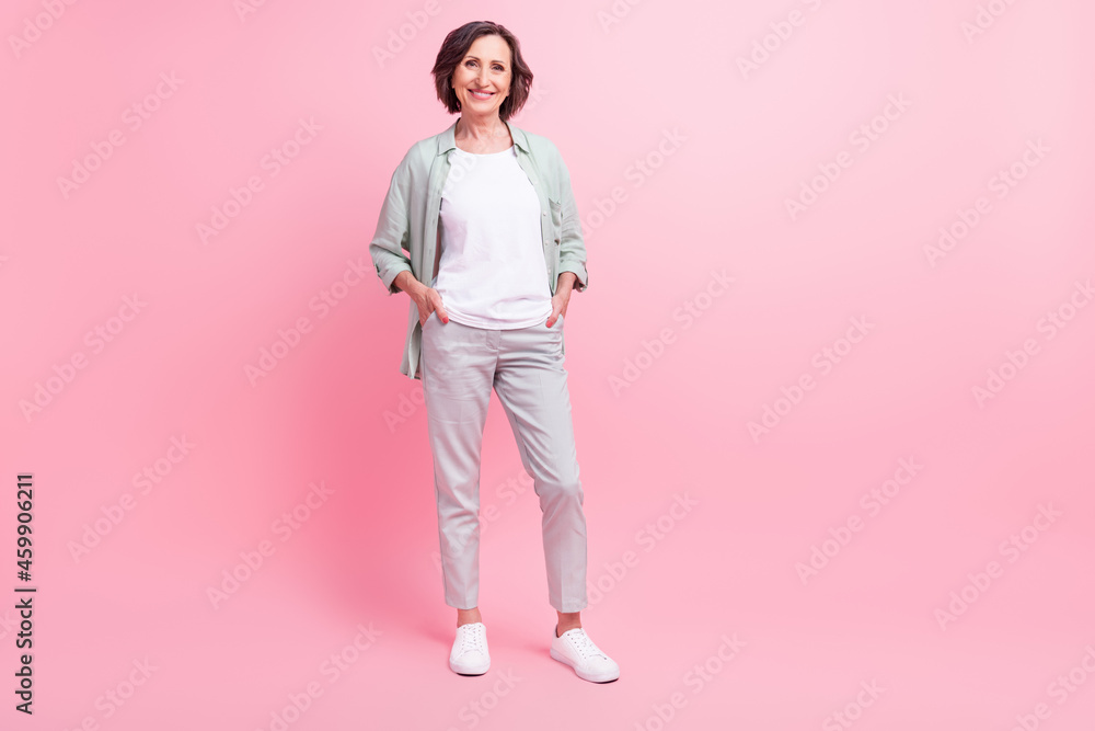 Full size portrait of cheerful pretty aged person put hands in pockets beaming smile isolated on pink color background Full size portrait of cheerful pretty aged person put hands in pockets beaming smile isolated on pink color background