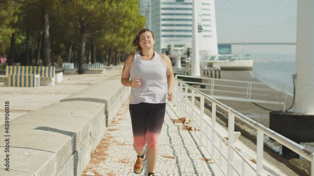 Long shot of cheerful body positive woman running in park. Front view of happy Caucasian woman in activewear jogging along path, doing cardio training. Outside activity, body positive, sport concept Long shot of cheerful body positive woman running in park. Front view of happy Caucasian woman in activewear jogging along path, doing cardio training. Outside activity, body positive, sport concept
