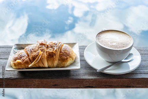 Latte art coffee in a white cup with croissant in white plate on on a wooden plank isolated blue sky reflection on lake
