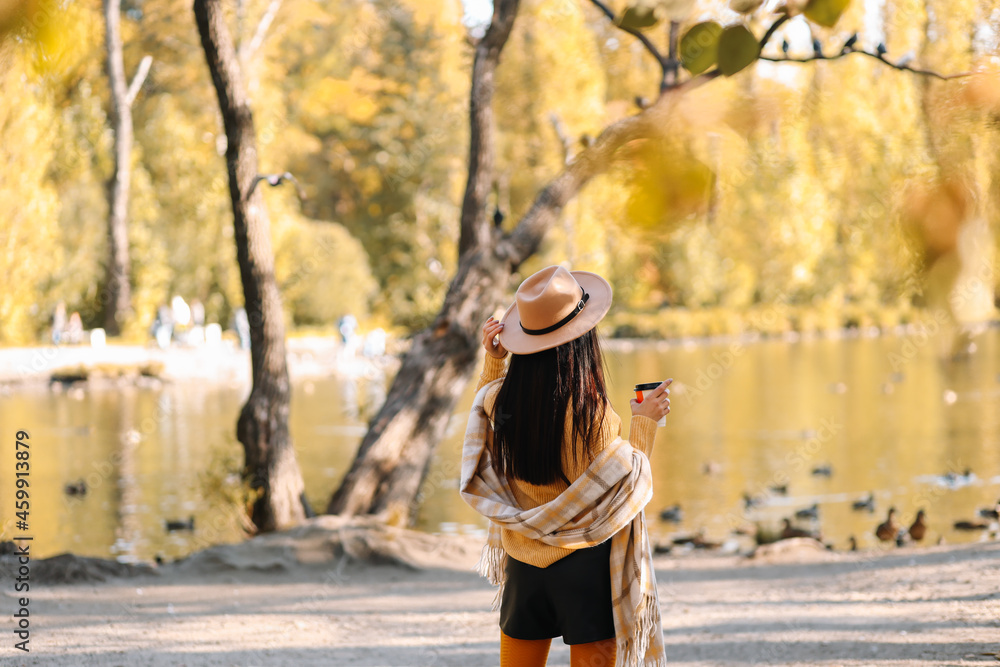 Portrait of a happy cheerful young woman in fashionable casual clothes and a stylish hat walking drinking coffee enjoying solitude in a fall park in nature by the water in autumn, selective focus