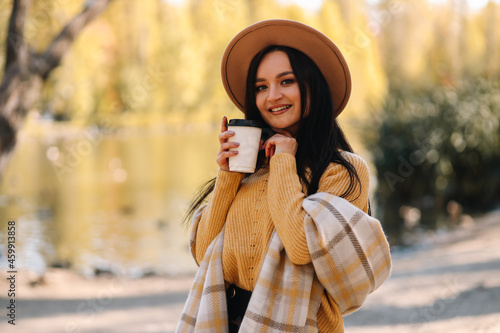 Portrait of a happy cheerful young woman in fashionable casual clothes and a stylish hat walking drinking coffee enjoying solitude in a fall park in nature by the water in autumn, selective focus © Елизавета Старкова