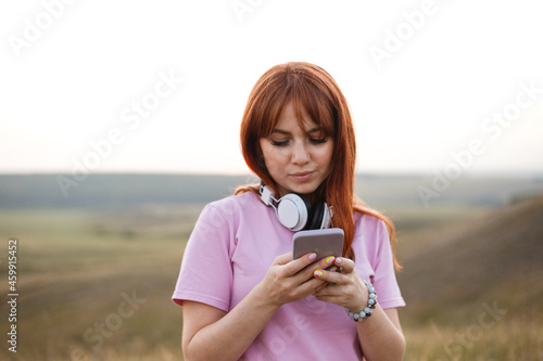 Young redhead woman with white headphones looks in the smartphone photo