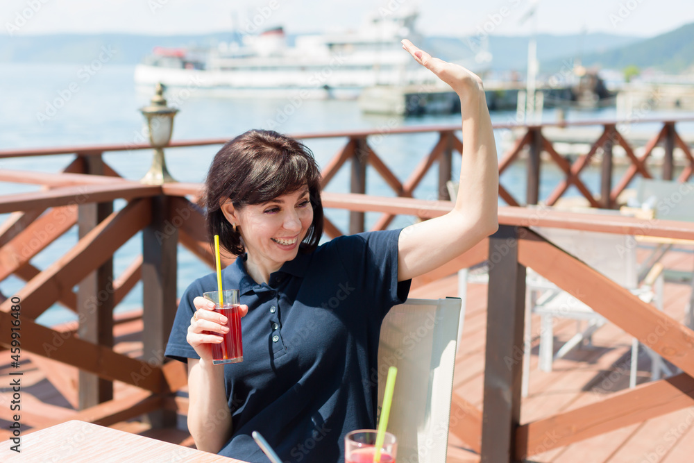 brunette in a cafe by the sea with a glass of juice happily waves to the waiter.