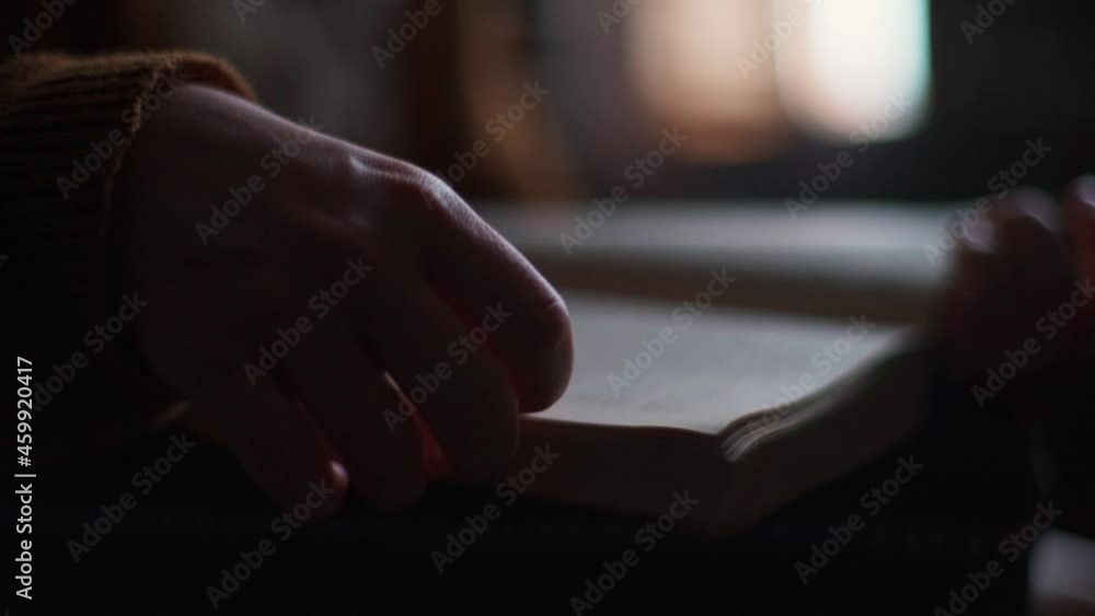 Close-up hands of unrecognizable young woman flipping through pages of old frayed book at home in evening. Side view of female hands turning pages of old book on blurred background of window. Close-up hands of unrecognizable young woman flipping through pages of old frayed book at home in evening. Side view of female hands turning pages of old book on blurred background of window.