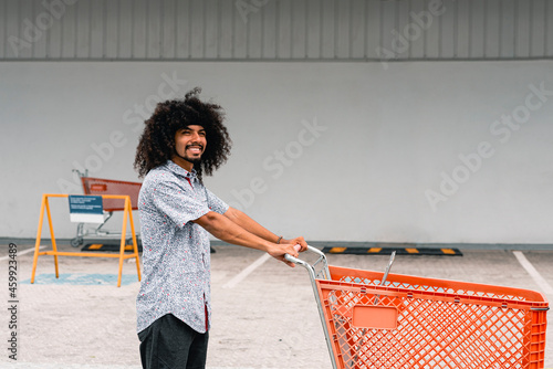 Obraz na plátně Hombre con cabello rizado muy sonriente con un carrito de compras en el parqueo