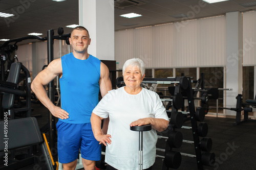 pensioner, an elderly woman performs an exercise with a barbell in gym. coach helps and shows. Healthy sports lifestyle, senior concept. woman with gray hair is recovering from injury. portrait