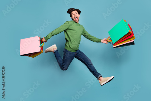 Full body photo of brunette happy amazed man jump run sale hold bags wear casual clothes isolated on blue color background