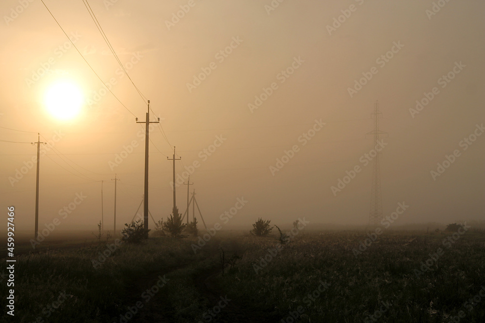 Power lines at sunrise. Power lines at sunrise. Country road at dawn in the fog
Power lines at dawn in the fog. Fog over the field