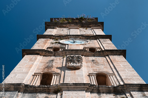 The tall bell tower is a symbol of Perast. A stone clock tower overgrown with grass and flowers. Bottom up view against clear blue sky. Catholic church of Saint Nicholas photo