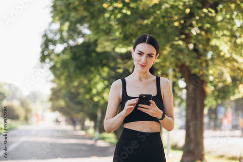 Stylish girl in black top posing on alley holding smartphone. Happy woman with beautiful smile chatting on cell smartphone while relaxing in the park, outdoors