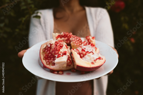 woman hands holding dish with fresh pomegranate pieces harvested from tree in the background with red seeds. concept of eating autumn fruit for healthy diet and lifestyle