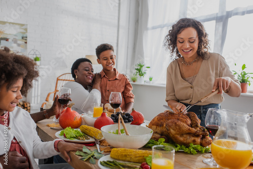 Smiling african american woman cutting turkey near family and thanksgiving dinner