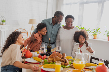 African american girl holding smartphone near family and thanksgiving dinner