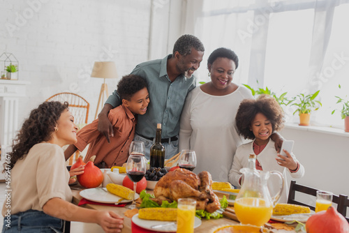 African american girl holding smartphone near family and thanksgiving dinner