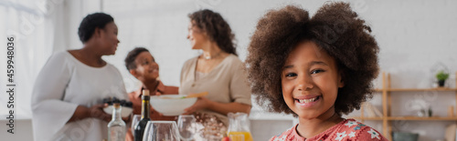 Smiling african american girl looking at camera near blurred family and thanksgiving dinner, banner
