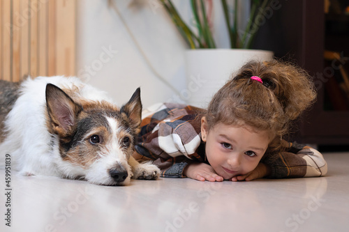 Little cute girl in a checkered dress and a dog are playing in the room, lie next to each other on the floor, friendship between a child and a pet, children and animals.