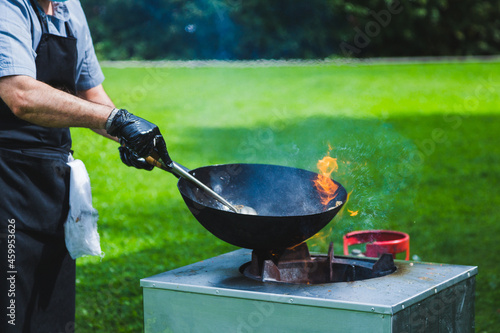 Chef prepares Chinese wok noodles over an open fire on a green lawn.