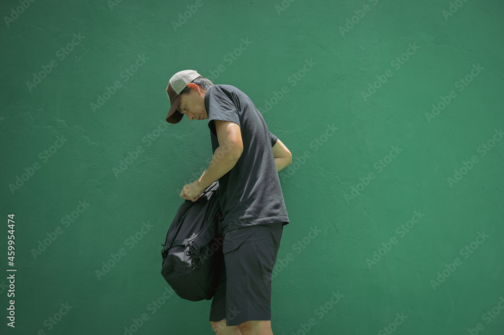 Side view point of young man wearing cap and carrying baggage standing in front of green color cement wall