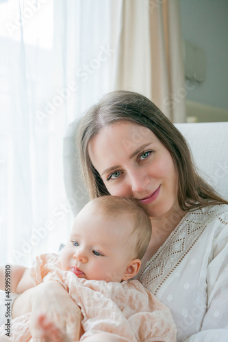 Happy young mother holds her little daughter 5 months old in her arms sitting in a chair at home. Motherhood.