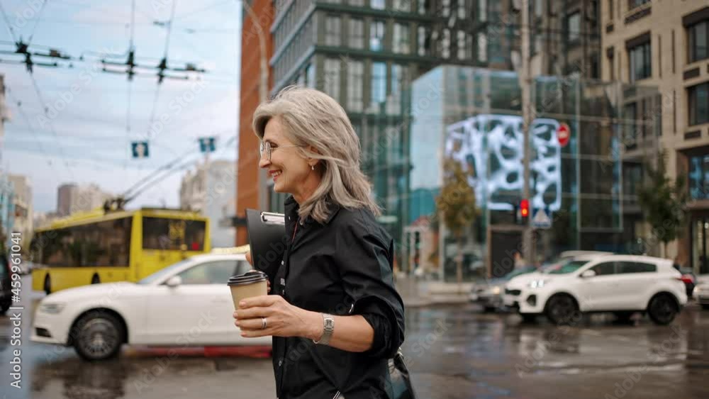 Adult gray-haired woman in glasses walks along a city street with a folder and coffee in her hands. Business lady walk. Slow motion Adult gray-haired woman in glasses walks along a city street with a folder and coffee in her hands. Business lady walk. Slow motion