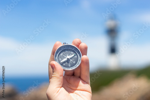 Hand holding a compass in front of a lighthouse by the sea, during a sunny day, concepts, selective focus.