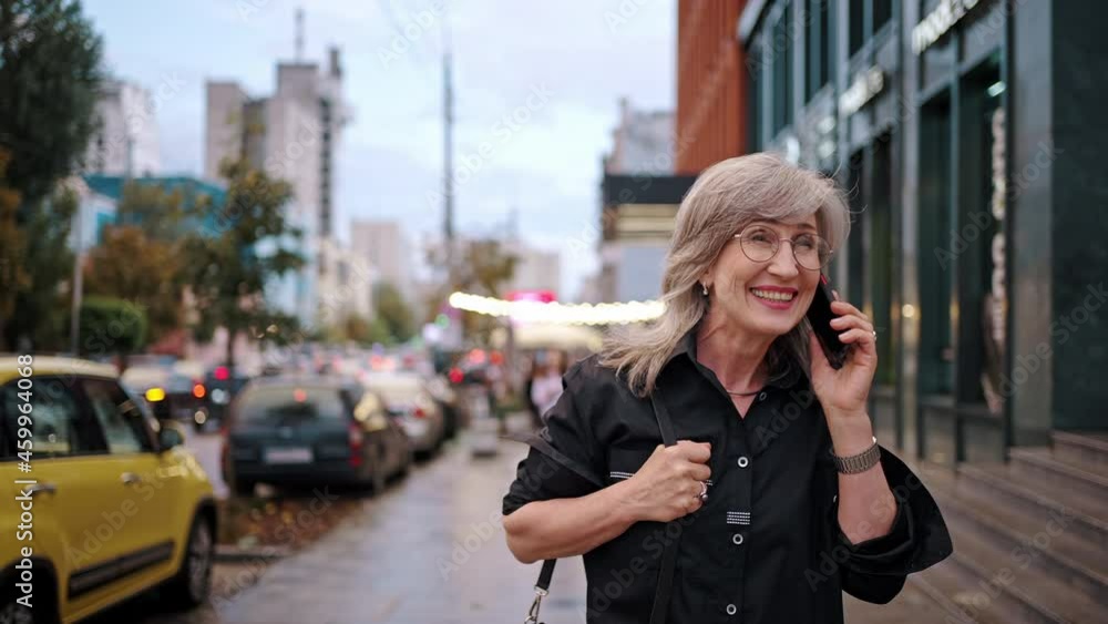 Happy good looking business woman walking along the city street and talking on the phone in the evening. Gray-haired lady wearing glasses walks with a smartphone in her hands. People and gadgets Happy good looking business woman walking along the city street and talking on the phone in the evening. Gray-haired lady wearing glasses walks with a smartphone in her hands. People and gadgets
