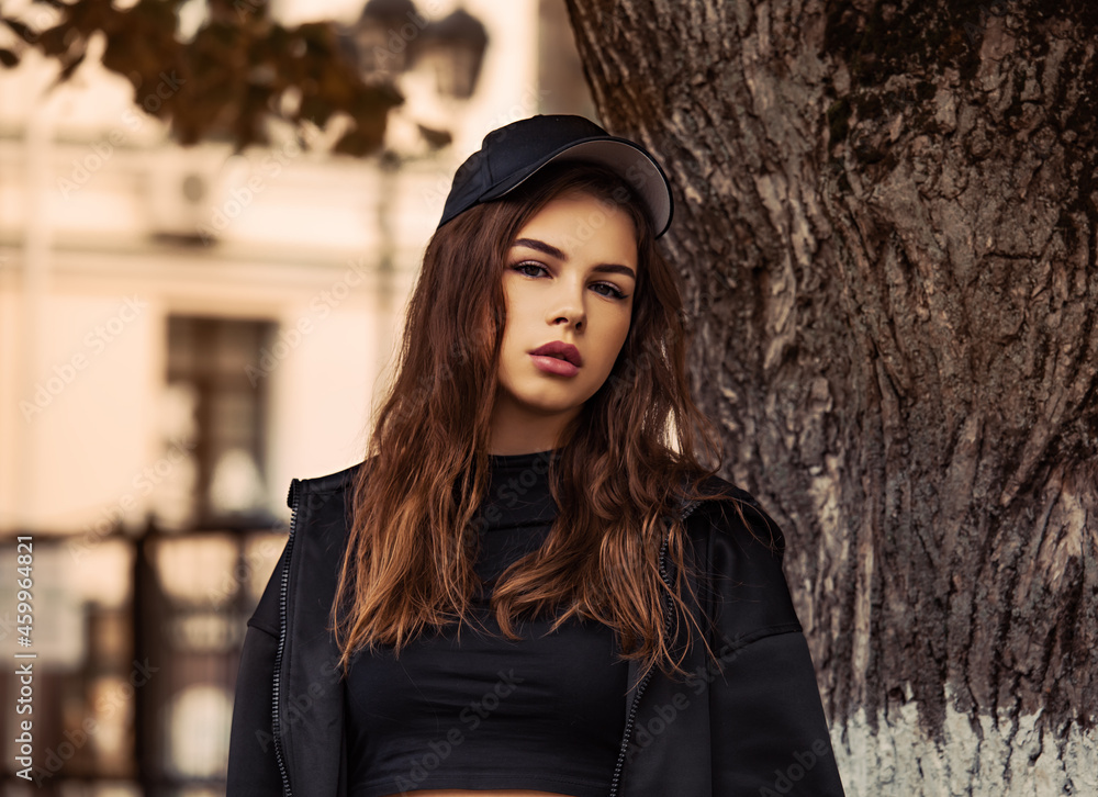 Young thinking teen woman looking near the tree  on the street background in black style clothing and cool cap. Autumn season clothing.