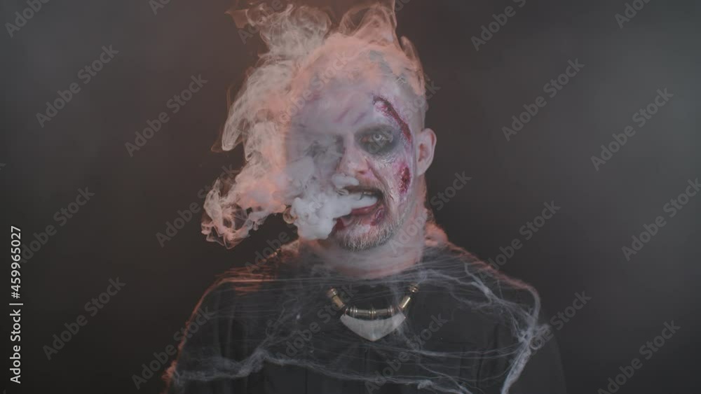 Sinister man with horrible scary Halloween zombie makeup in costume looking ominous at camera blows smoke from nose and mouth. Dead guy with wounded bloody scars face isolated against black background