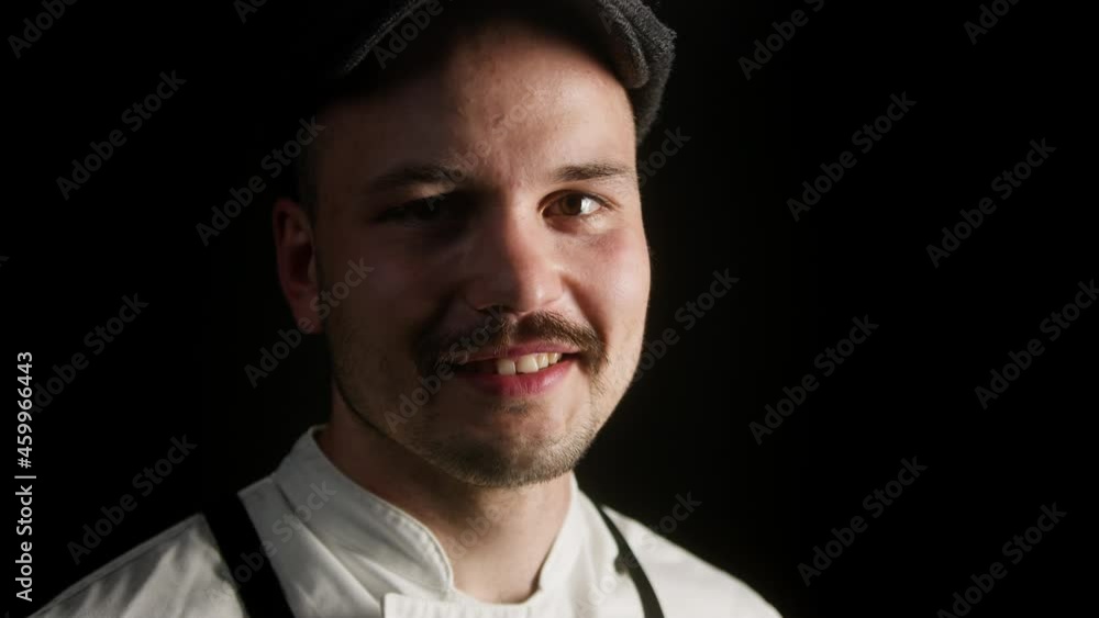 Professional chef standing on black background. Portrait of smiling man with moustache wearing kitchen uniform, apron. Handsome cheerful cook looking in camera.