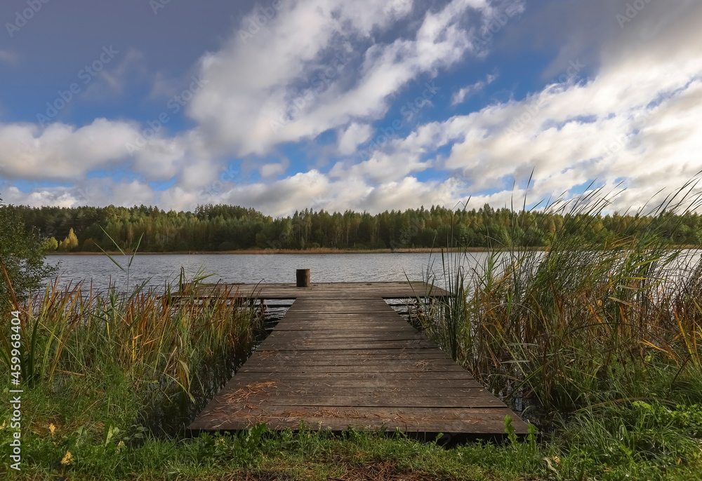 Obraz premium Landscape with wooden long jetty with chair for fishing, lake, forest on horizon and cloudy blue sky in summer.