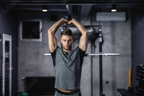 Pumping arm and shoulder. A young attractive man in a gray shirt raises dumbbell above his head in the gym. Strengthening triceps and biceps. Initial body posture and sports discipline
