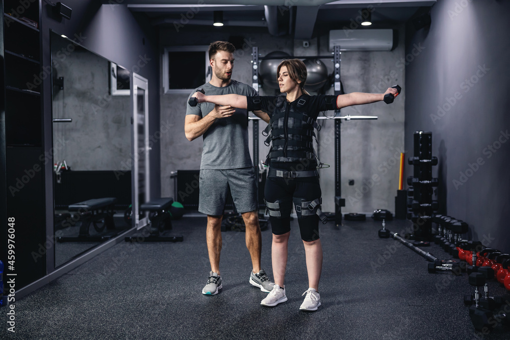 A revolution in training, a new technological approach to training. A male instructor assists the women in a special suit for EMS technology while lifting dumbbells with both hands in a modern gym