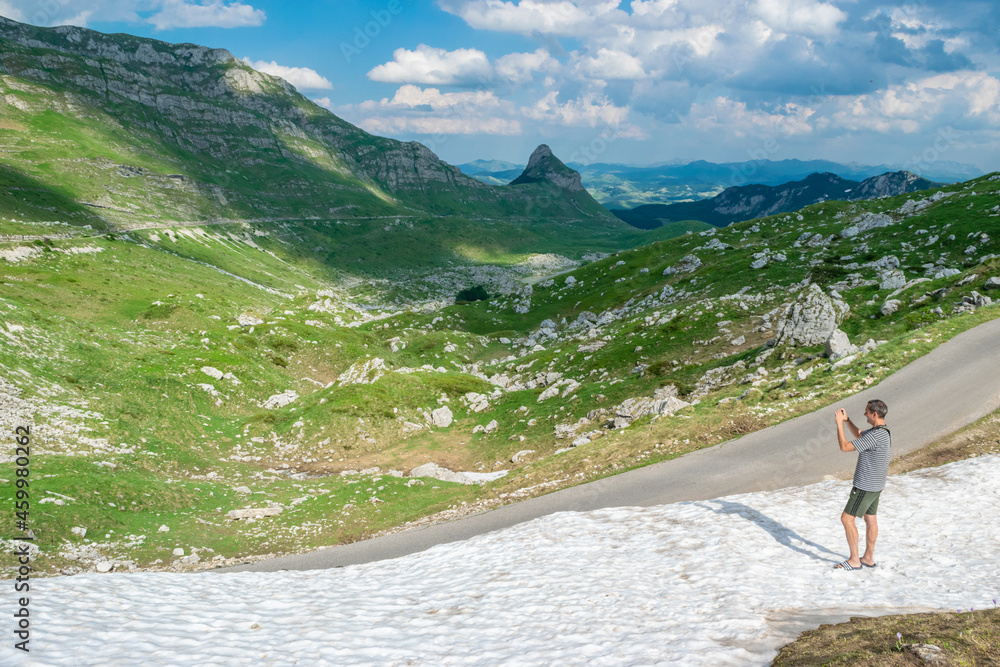 Tourist man takes a photo of beautiful summer landscape while standing in snow. Durmitor National Park. Highest road pass in Montenegro.