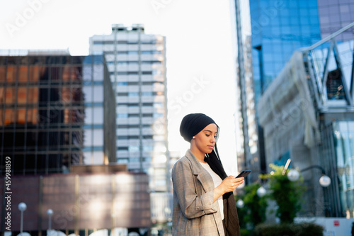 Side view of an adult muslim woman standing in a urban area using her phone