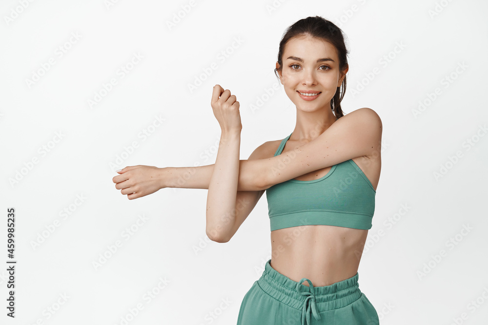 Smiling fit fitness girl stretching arms. Female athlete doing workout exercises, warm-up before training session in gym, white background Smiling fit fitness girl stretching arms. Female athlete doing workout exercises, warm-up before training session in gym, white background