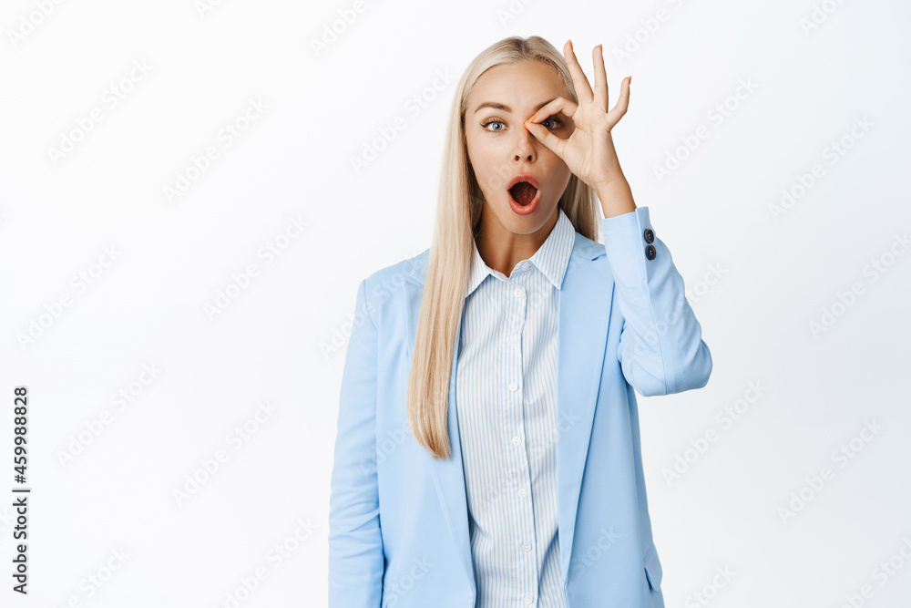 Image of saleswoman showing zero, ok gesture, looking through okay sign with amazed face expression, standing in suit over white background