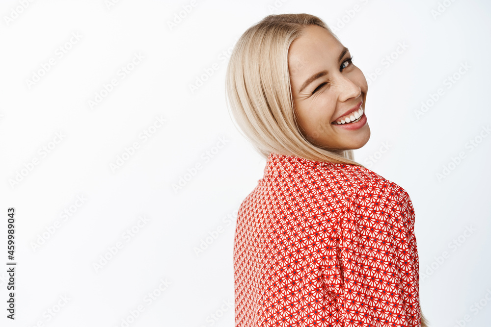 Women beauty. Feminine blond girl turn around, looking behind her shoulder and winking coquettish at camera, standing over white background Women beauty. Feminine blond girl turn around, looking behind her shoulder and winking coquettish at camera, standing over white background