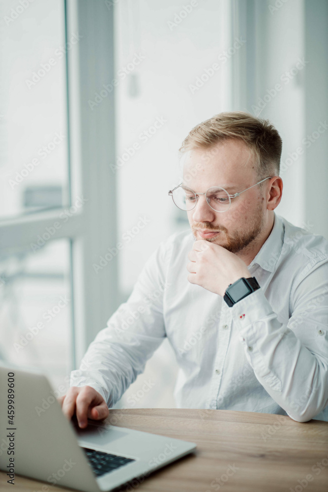 young caucasian man working with a laptop in a light office wearing glasses, the concept of working on the internet of a young specialist. young caucasian man working with a laptop in a light office wearing glasses, the concept of working on the internet of a young specialist.