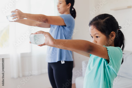 Asian young mother and her daughter doing stretching fitness exercise yoga and using the bottle of water to be dumbbells together at home in daily routine.