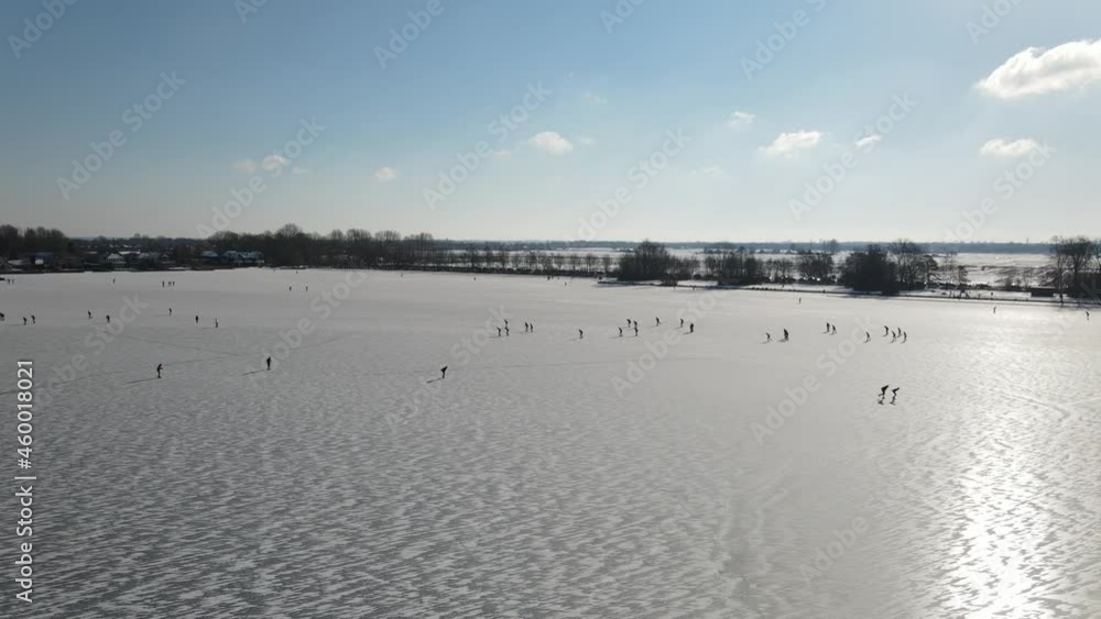 Aerial drone shot over frozen lake during cold winter day with kids ice skating during sunlight
