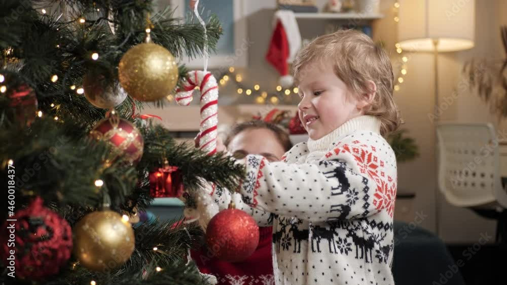 Mother and kid are decorating Christmas tree. Mom and child of 2-3 years old at home decorate Christmas tree, put toys on branches. Slow motion Mother and kid are decorating Christmas tree. Mom and child of 2-3 years old at home decorate Christmas tree, put toys on branches. Slow motion