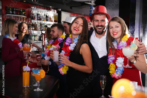 Cheerful guy with two girls dancing and toasting drinks on Hawaiian party in bar