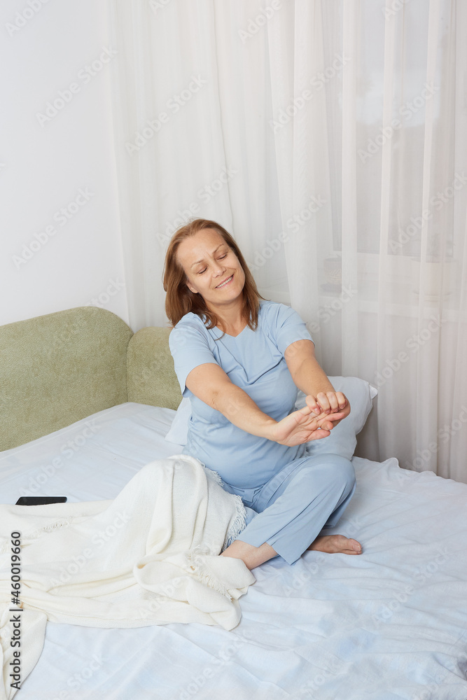 Mature happy woman in blue pajamas stretching in the morning after sleep while sitting in bed