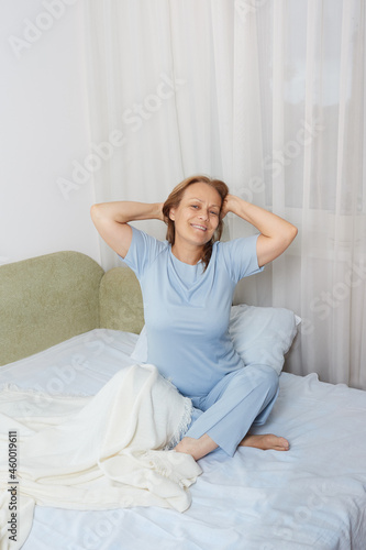 Calm smiling senior woman in blue pajamas stretching in the morning after sleeping in her bed