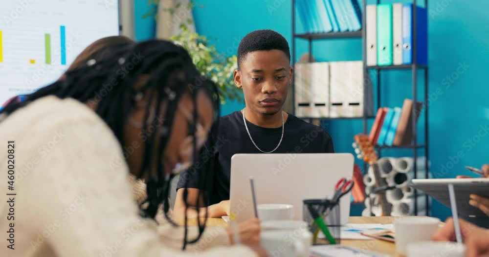 Boy working in office for large marketing firm sits at table with co-workers taking notes, making charts, schedules, man enters data into system on laptop Boy working in office for large marketing firm sits at table with co-workers taking notes, making charts, schedules, man enters data into system on laptop