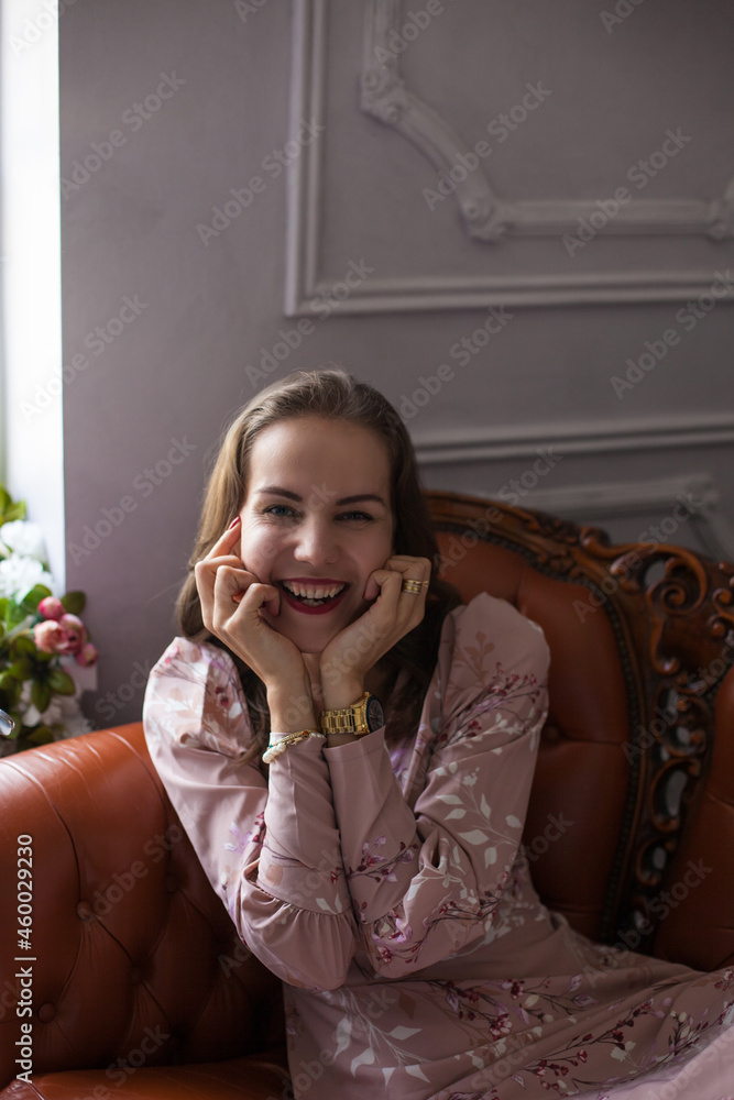 Fototapeta premium Young beautiful woman in pink dress and stylish accessories sits on brown leather sofa on white wall background. Portrait of happy smiling girl with red lips resting at home and looking at camera. 