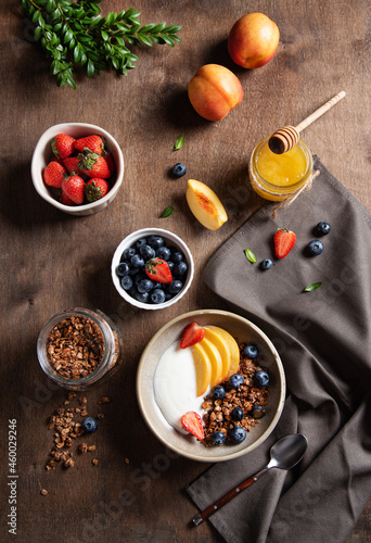 Concept of a healthy and nutritious breakfast. Delicious natural yogurt with homemade granola, peach, strawberry, honey and blueberry in a bowl on a dark wooden background. Diet food. Top view image