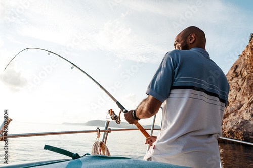 Young african american man standing with fishing rod on a sailboat fishing in open sea on sunset photo